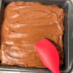 Brownies with chocolate frosting on top. There's a red spatula spreading the frosting.