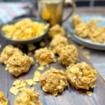 Corn Flake cookies on a wooden cutting board. There's a bowl of Corn Flakes and a cup of coffee next to the cookies.