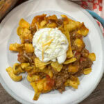 Frito pie on a white plate. There's some sour cream on top and a few sprinkles of Mexican shredded cheese.