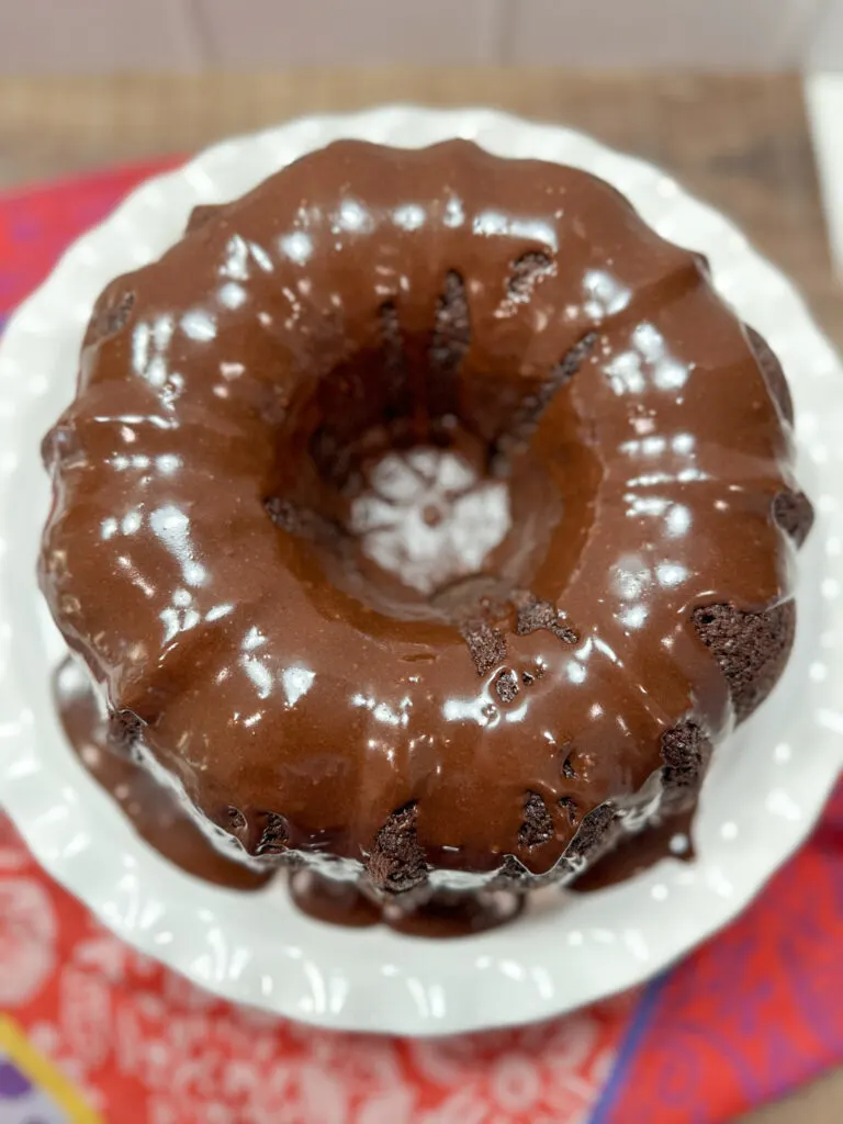 Chocolate cake with frosting on a white cake stand. There's a red kitchen towel below the stand.