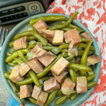 Cooked green beans and ham on a blue plate. The plate is sitting in front of a slow cooker. There is a red, white, and teal towel underneath the plate.