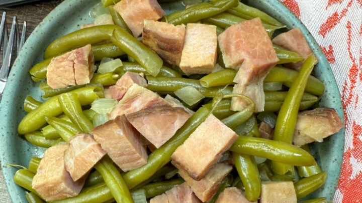 Cooked green beans and ham on a blue plate. The plate is sitting in front of a slow cooker. There is a red, white, and teal towel underneath the plate.