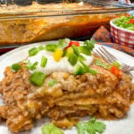 A piece of enchilada casserole topped with sour cream and diced bell peppers. The casserole dish is in the background, as well as a bowl of diced green, yellow, and red bell peppers.