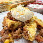 Mexican cornbread casserole with ground beef on a white plate. The casserole is topped with sour cream and shredded cheese. The casserole dish is in the background along with a bowl of salsa and sour cream.