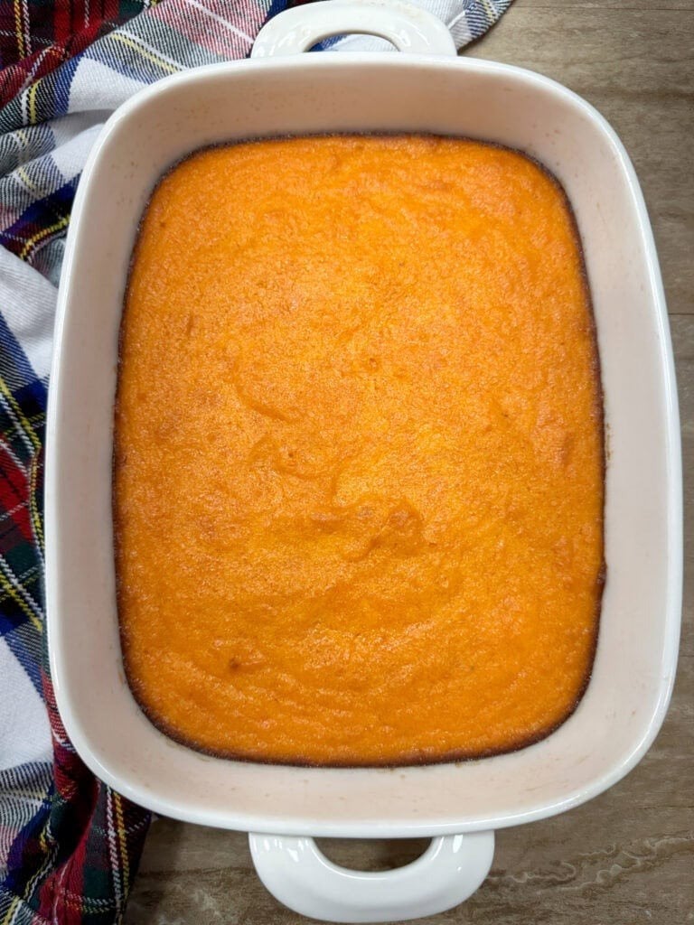 A white rectangular baking dish filled with smooth, orange casserole—just like How To Make Copycat Ruths Chris Sweet Potato Casserole—sits on a wooden surface next to a colorful plaid cloth, its edges slightly browned.