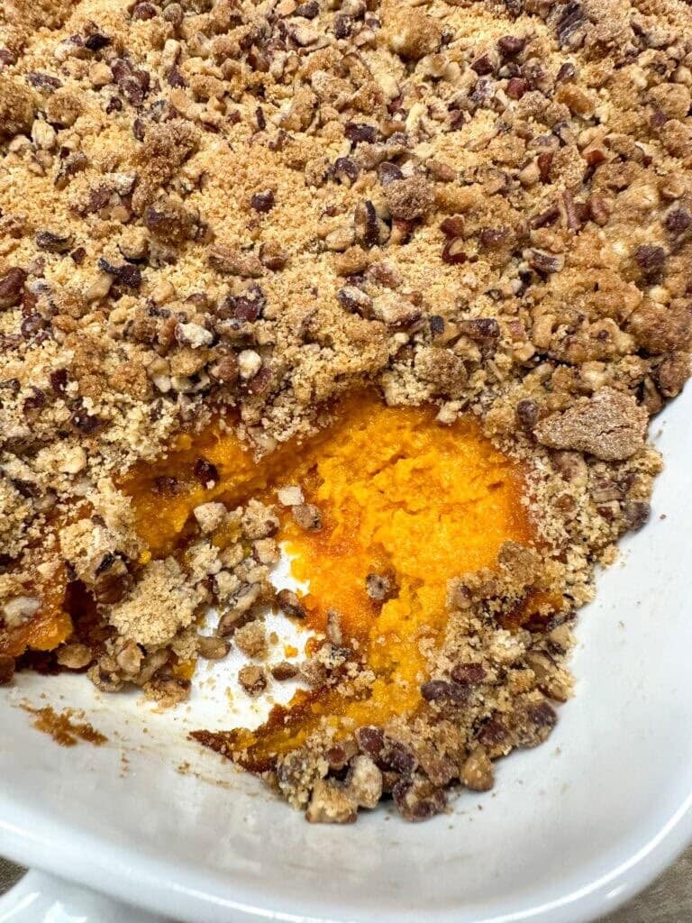 A close-up of a sweet potato casserole topped with a crumbly pecan and brown sugar streusel, with a scoop missing to reveal the bright orange filling—just like you’ll find in How To Make Copycat Ruths Chris Sweet Potato Casserole.
