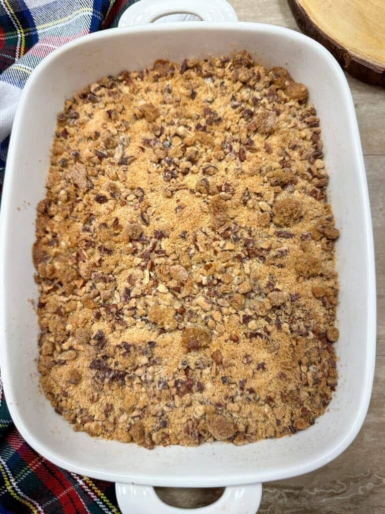 A white rectangular baking dish filled with a golden-brown, crumbly dessert topped with chopped nuts sits on a light surface next to a plaid cloth—just like in guides on How To Make Copycat Ruths Chris Sweet Potato Casserole.