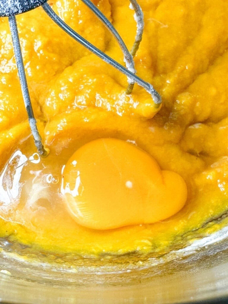 A close-up of a raw egg yolk and egg white atop yellow batter in a mixing bowl, with metal beaters partially visible above the mixture—capturing an early step in How To Make Copycat Ruths Chris Sweet Potato Casserole.