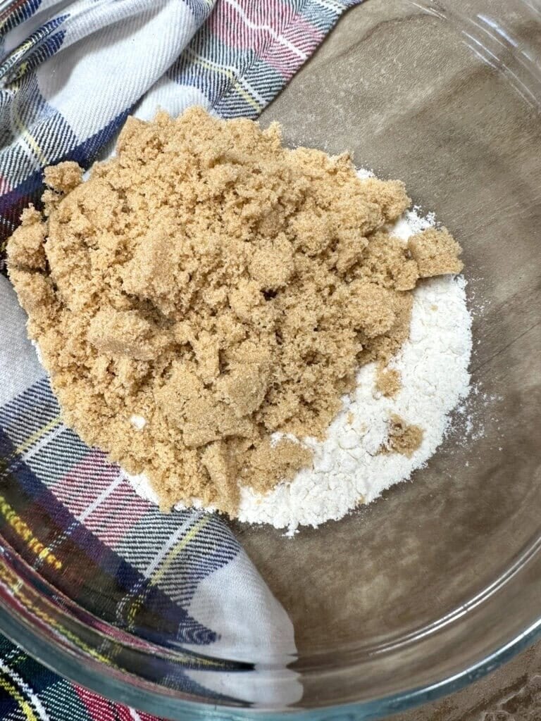 A glass bowl with a mixture of brown sugar and flour sits on a wooden surface, partially covered by a colorful plaid cloth—just one step in learning How To Make Copycat Ruths Chris Sweet Potato Casserole.