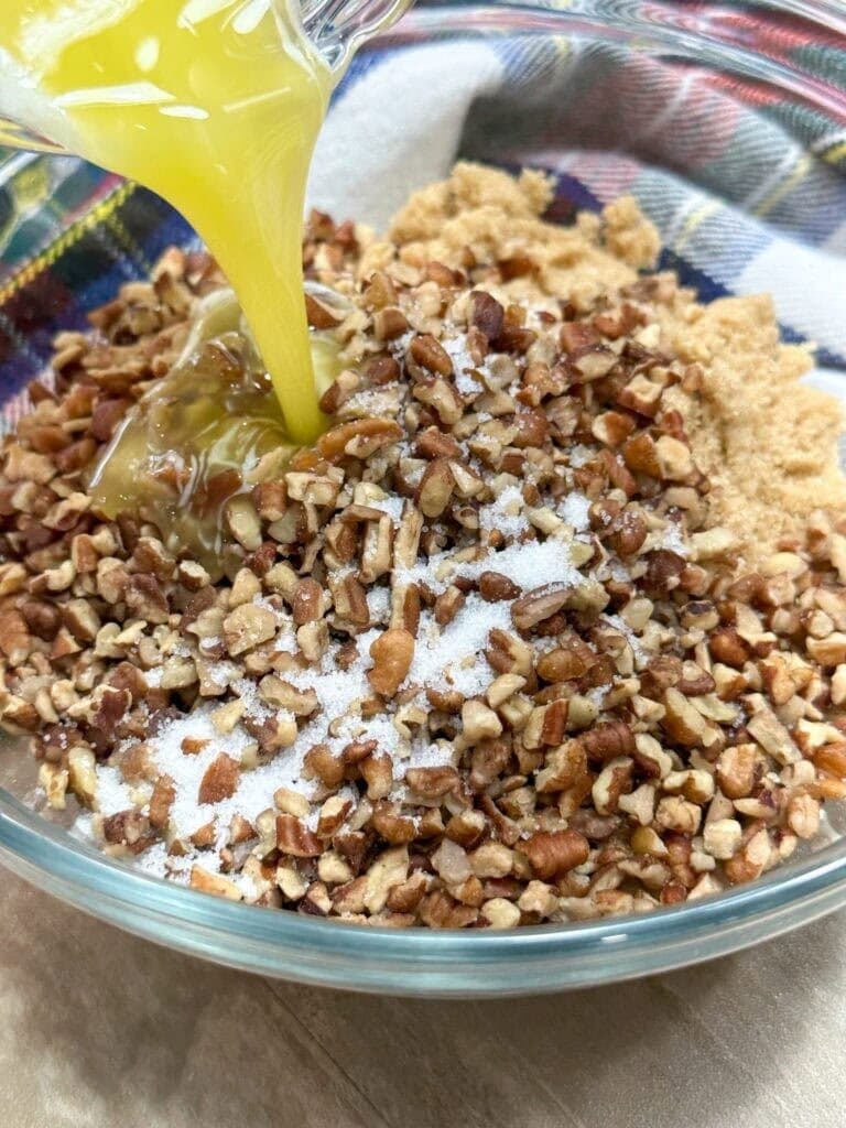 Chopped pecans, brown sugar, and salt in a glass bowl, with melted butter being poured in—this is the crunchy topping step in How To Make Copycat Ruths Chris Sweet Potato Casserole, set against a colorful cloth background.