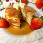 A stack of pancakes on a white plate with strawberries on the plate.