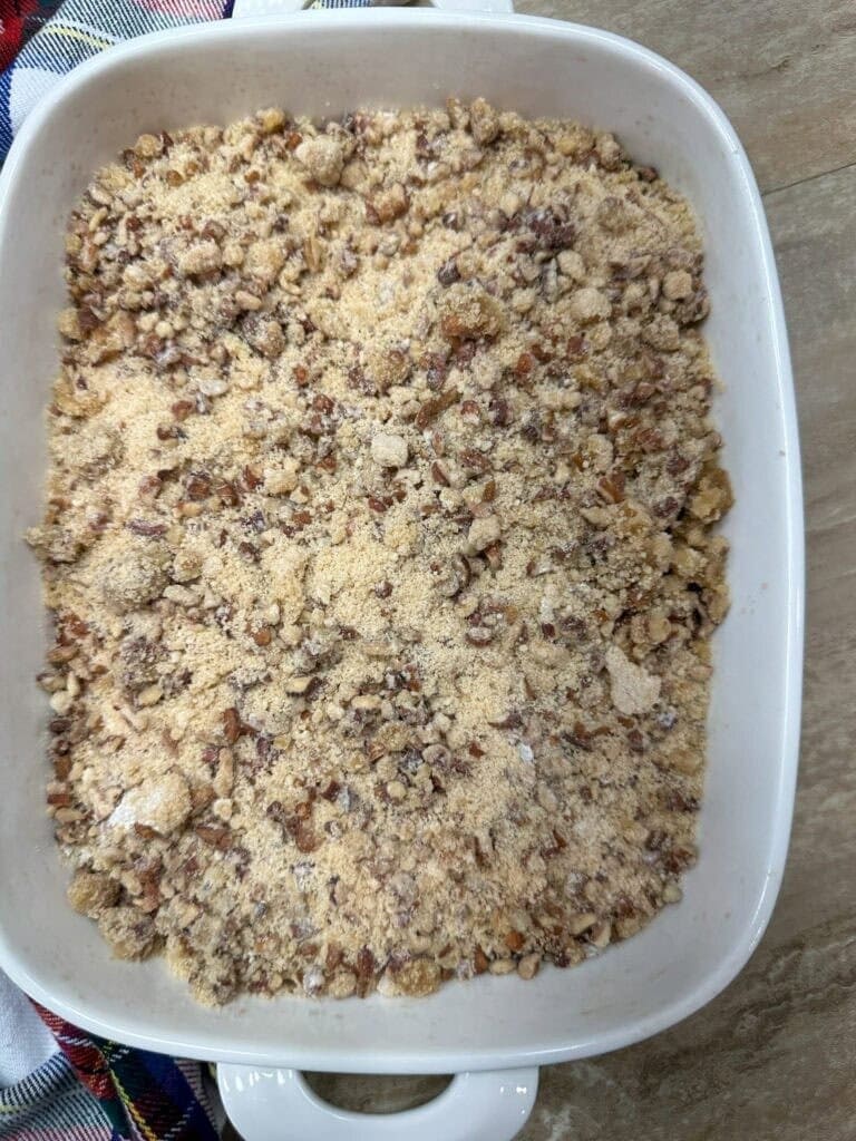 A white baking dish filled with an unbaked crumbly mixture of brown sugar, nuts, and flour sits on a wooden surface next to a plaid cloth—just the start of How To Make Copycat Ruths Chris Sweet Potato Casserole.