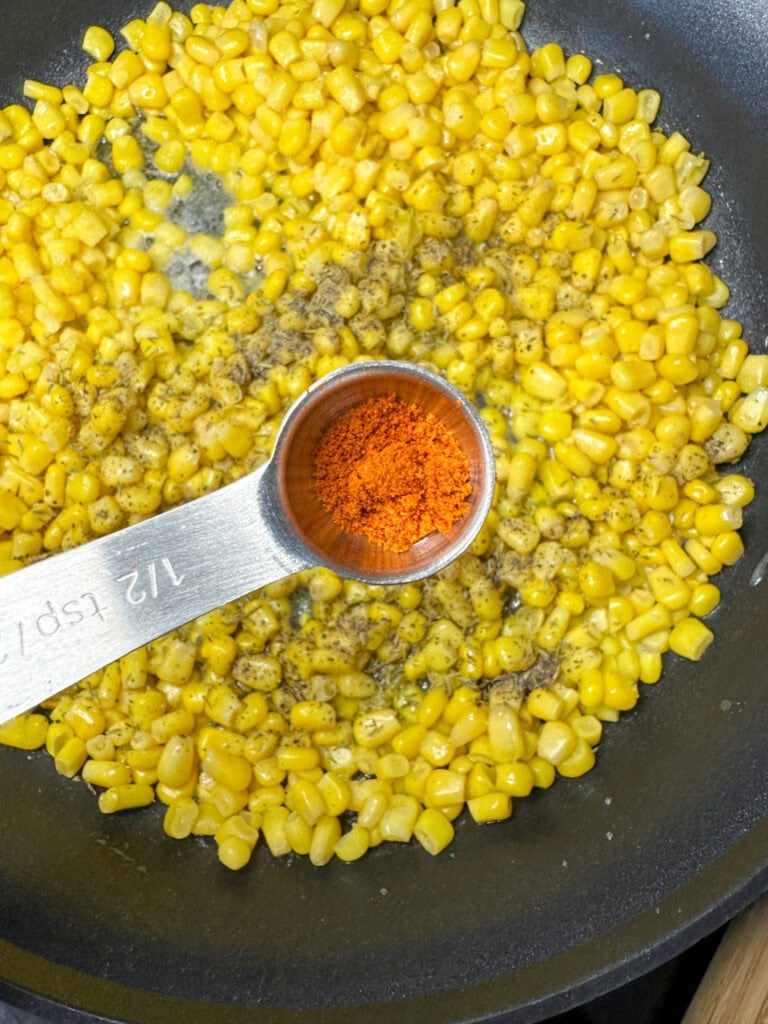 Corn in a skillet with a teaspoon filled with seasoning.