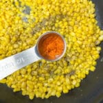 Corn in a skillet with a teaspoon filled with seasoning.