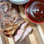 grilled pork steak on a cutting board. There are two slices in the pork. There's some broccoli and barbecue sauce in two different bowls next to the pork.