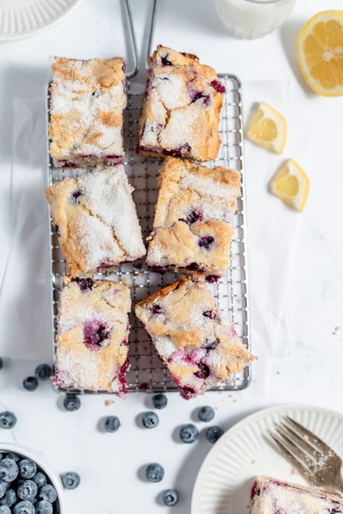 Six slices of coffee cake on a cooling rack. There are lemons in the background as well as blueberries in a bowl and scattered on the counter. 