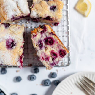Four slices of lemon blueberry coffee cake on a cooling rack. There are blueberries and lemon slices on the counter.