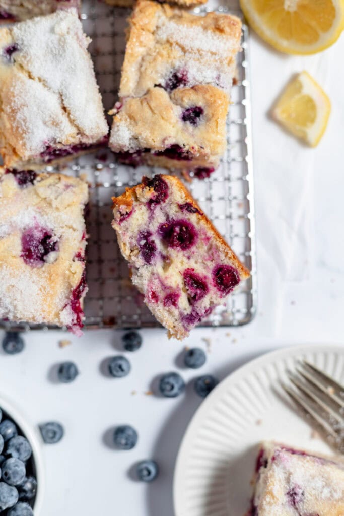 Four slices of lemon blueberry coffee cake on a cooling rack. There are blueberries and lemon slices on the counter. 