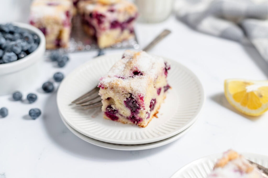 A slice of lemon blueberry coffee cake on two white plates. The plates are stacked on top of each other. There are some blueberries in a bowl next to the plates and lemon on the counter. 