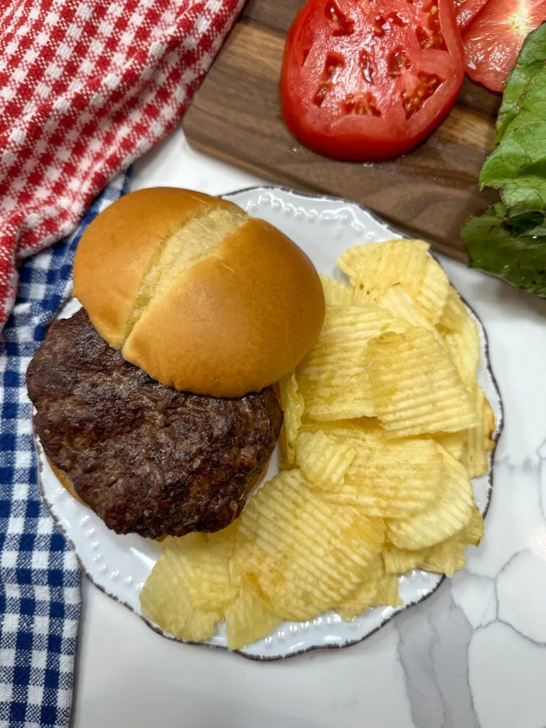 An air fryer burger on a bun with tomatoes and lettuce. There's some Ruffles chips on the white plate with the burger.