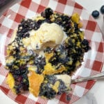 Blueberry dump cake on a red and white gingham plate. There's a fork with a bite sitting on the plate.