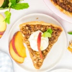 A slice of peach crisp pie on a white plate. There are two peach slices on the plate.