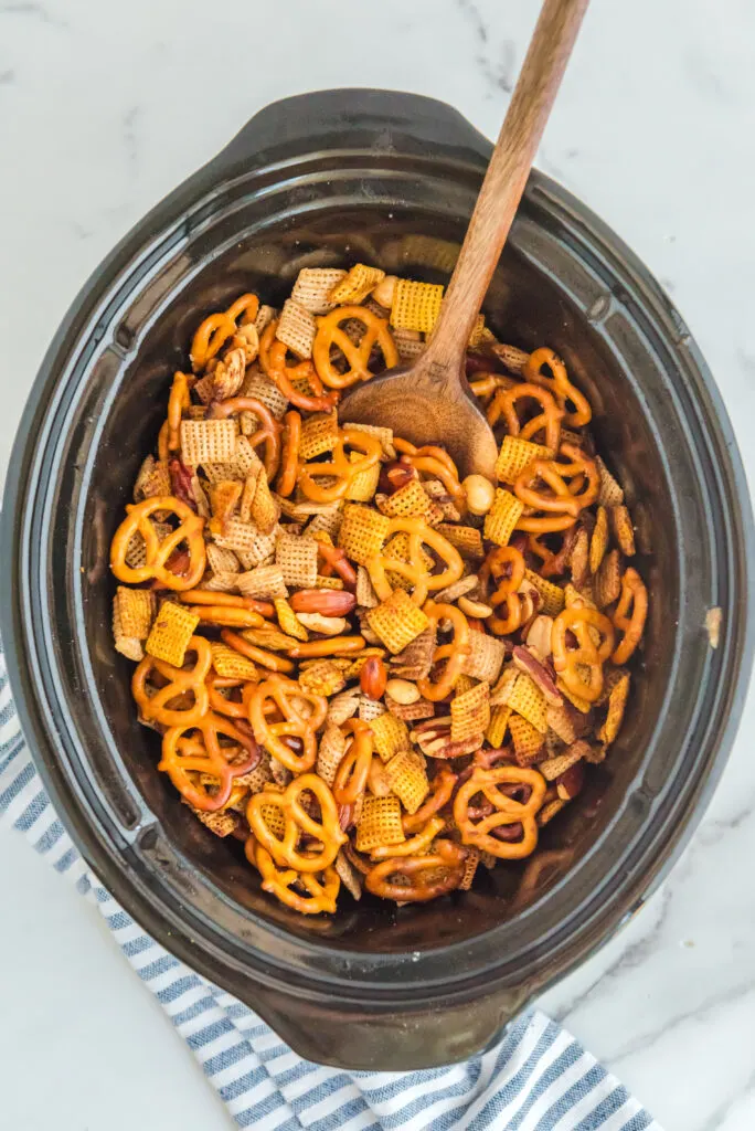 Chex Mix being stirred by a wooden spoon in a slow cooker.