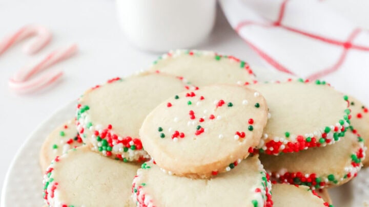 Sliced sugar cookies stacked on a white plate. There is a glass of milk in the background.