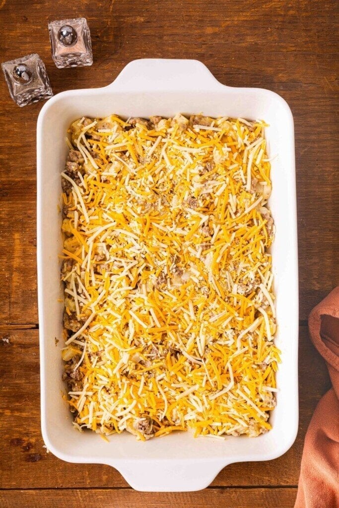 A white baking dish filled with 5-Ingredient Ground Beef Casserole, topped with shredded cheddar and mozzarella cheese, sits on a wooden table next to two square salt and pepper shakers and an orange cloth napkin.