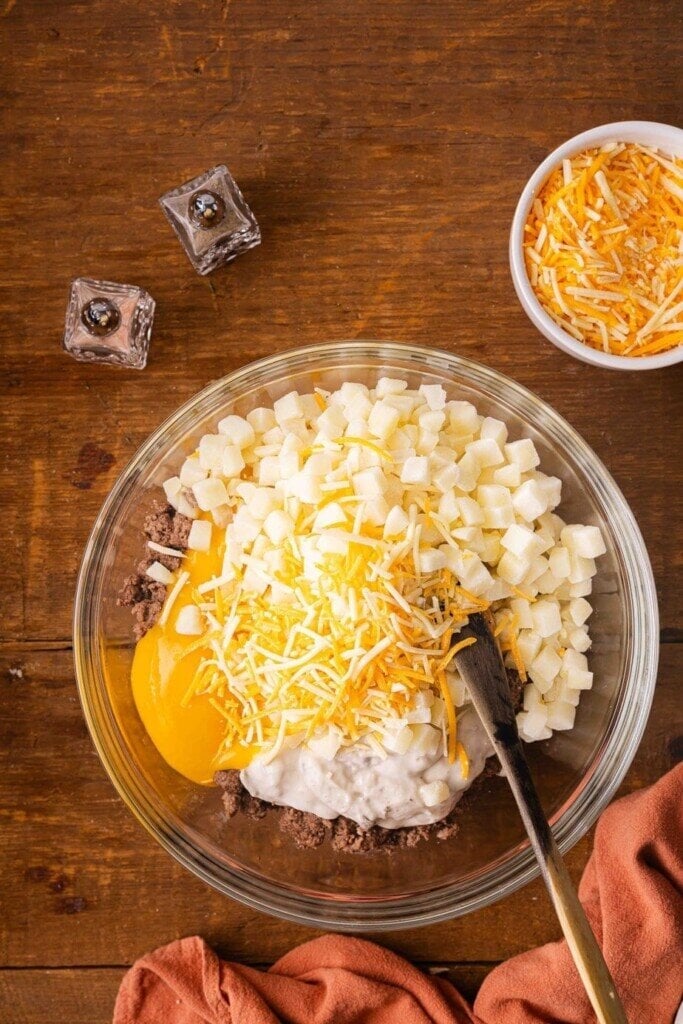 A glass bowl on a wooden table holds the makings of a 5-Ingredient Ground Beef Casseroleโdiced potatoes, shredded cheese, ground meat, cream of soup, and sour cream. A spoon rests inside, with toppings and seasonings nearby beside an orange cloth.