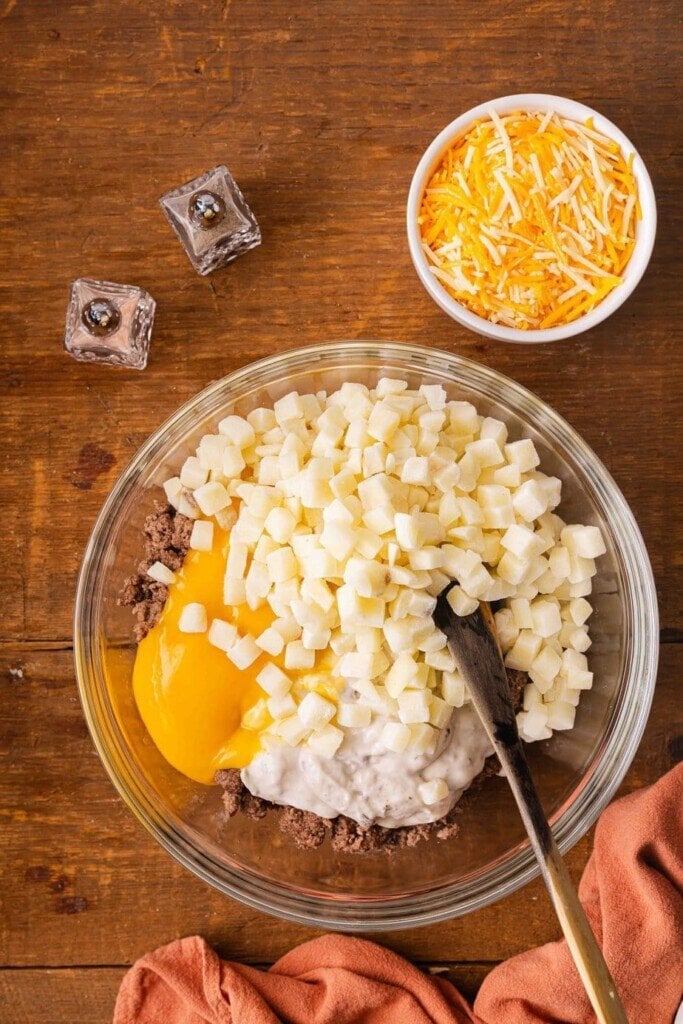 A glass bowl with 5-Ingredient Ground Beef Casseroleโground beef, cream of soup, sour cream, cubed potatoes, and shredded cheeseโsits on a wooden table with a spoon inside. A bowl of shredded cheese and salt and pepper shakers are nearby.