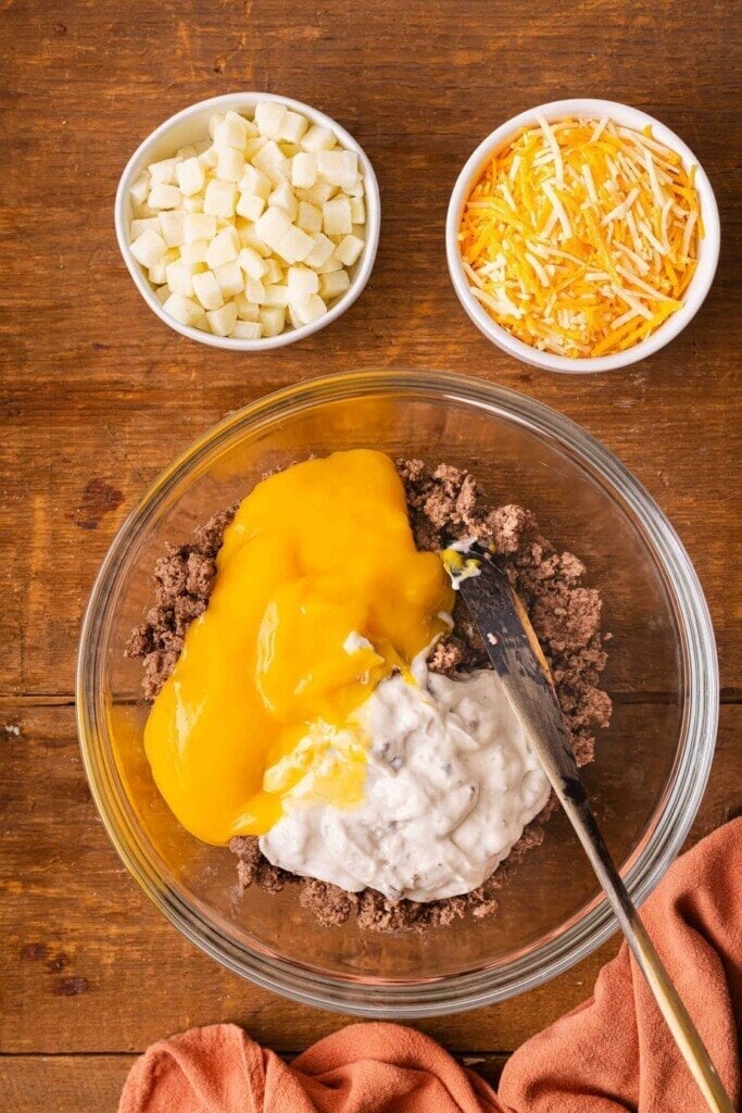 A glass bowl holds a 5-Ingredient Ground Beef Casserole with creamy soup and melted cheese on a wooden table. Nearby, small bowls are filled with cubed potatoes and shredded cheese, while a wooden spoon rests in the mixture.