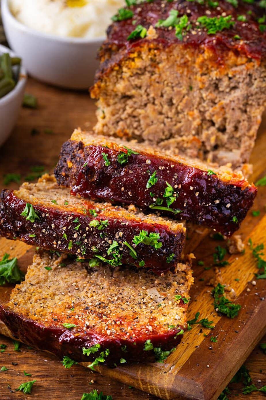 Close-up of a sliced meatloaf topped with ketchup glaze and fresh parsley on a wooden cutting board, with mashed potatoes in the background.
