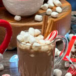 Hot chocolate in a glass with marshmallows, cinnamon sticks, and a candy cane.
