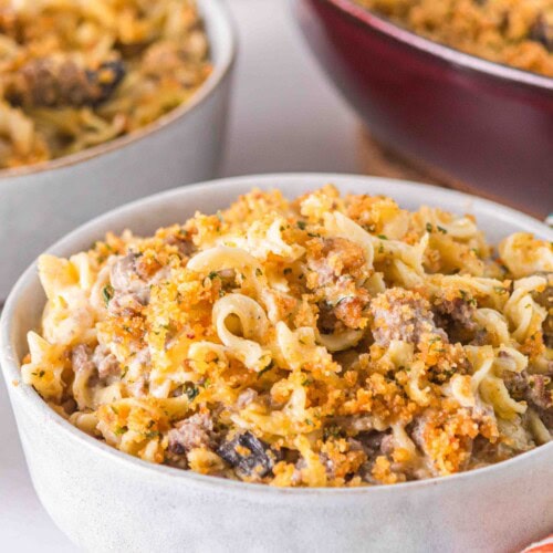A bowl of creamy One Pot Ground Beef Stroganoff Casserole topped with golden breadcrumbs sits on a table, with a colorful napkin nearby and more casserole in the background.