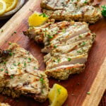 Grilled chicken breasts sliced and garnished with chopped parsley and lemon wedges on a wooden serving board. A small bowl of fresh parsley and lemon slices are in the background. Brown cloth napkins accent the setting.
