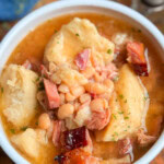 A bowl of navy beans with chunks of ham and pieces of bread dumplings. The dish is garnished with herbs. In the background, there are two salt and pepper shakers on a wooden surface.