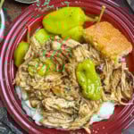 Red plate with shredded pork chops over mashed potatoes, garnished with green peppers and herbs. A square piece of cornbread is on the side. A spoon and wooden table background complete the rustic setting.