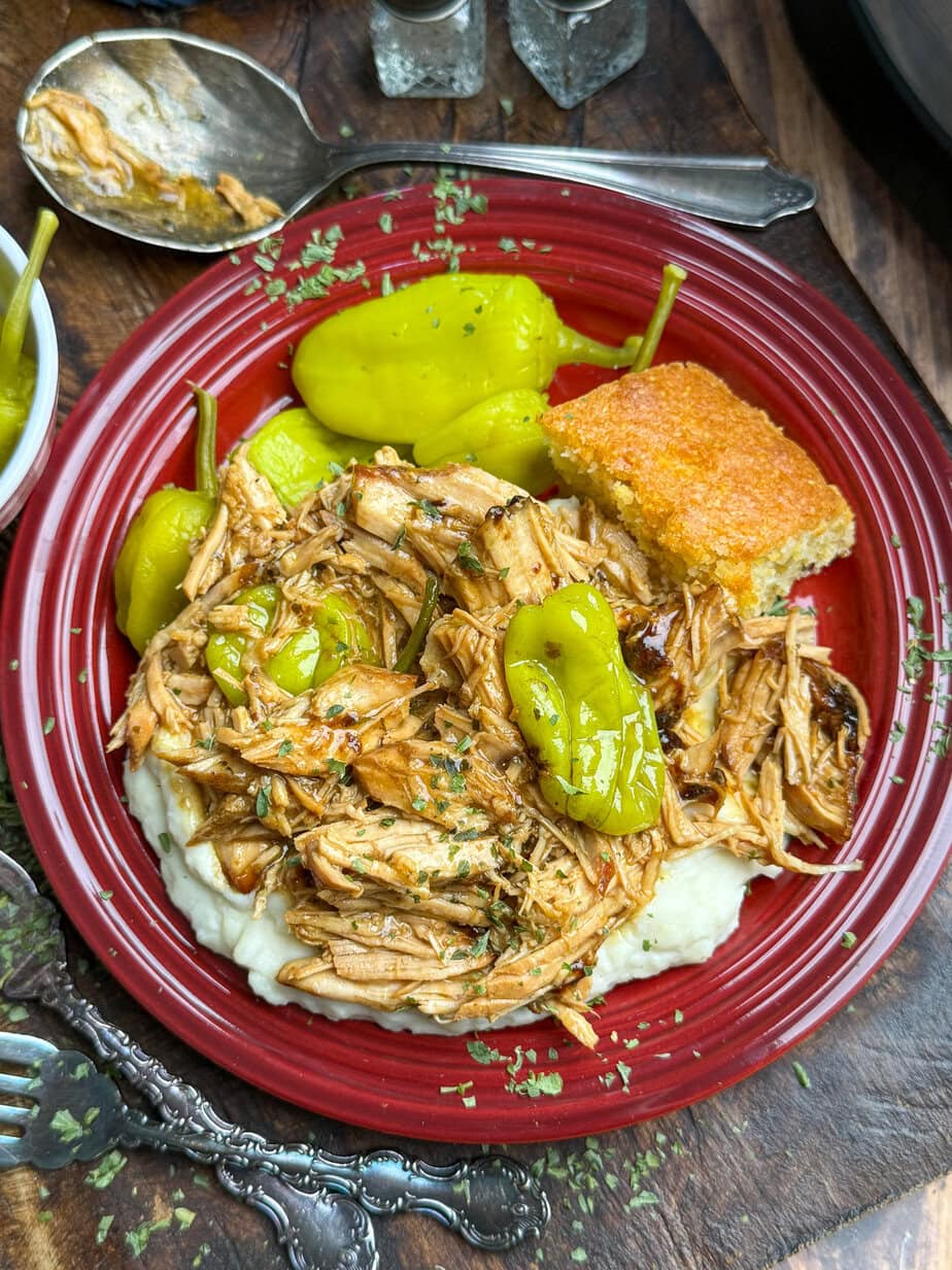 A red plate with shredded pork chops topped with pepperoncini peppers on a bed of mashed potatoes, accompanied by a piece of cornbread. A spoon with sauce and a bowl containing more peppers are visible in the background on a wooden surface.