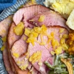 Slices of Ham topped with pineapple chunks resting on a plate, accompanied by green bean casserole and a buttered bread roll. Forks and a blue napkin complete the setting on the wooden table.