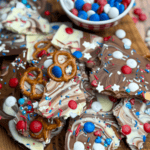 A wooden board topped with chocolate bark pieces decorated with red, white, and blue candies, sprinkles, and pretzels. A bowl of matching candies sits in the background. Text reads “PATRIOTIC CHOCOLATE BARK WITH CANDY.”.