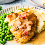 A plate with creamy mashed potatoes, green peas, and pork chops topped with a creamy bacon sauce. Another plate of food and a bowl of mashed potatoes are in the background.