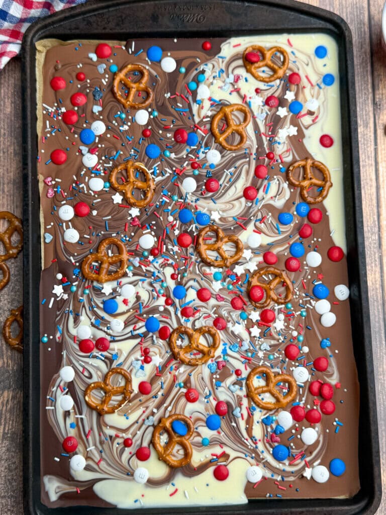 A baking tray with swirled white and milk chocolate bark, topped with pretzels, red, white, and blue candy-coated chocolates, star-shaped sprinkles, and red, white, and blue sprinkles.