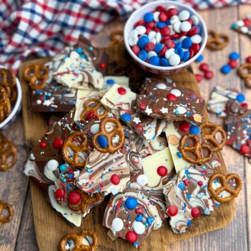 A wooden board topped with swirled chocolate bark pieces decorated with red, white, and blue candies, pretzels, and sprinkles. A bowl of matching candies sits nearby, with a plaid cloth in the background.