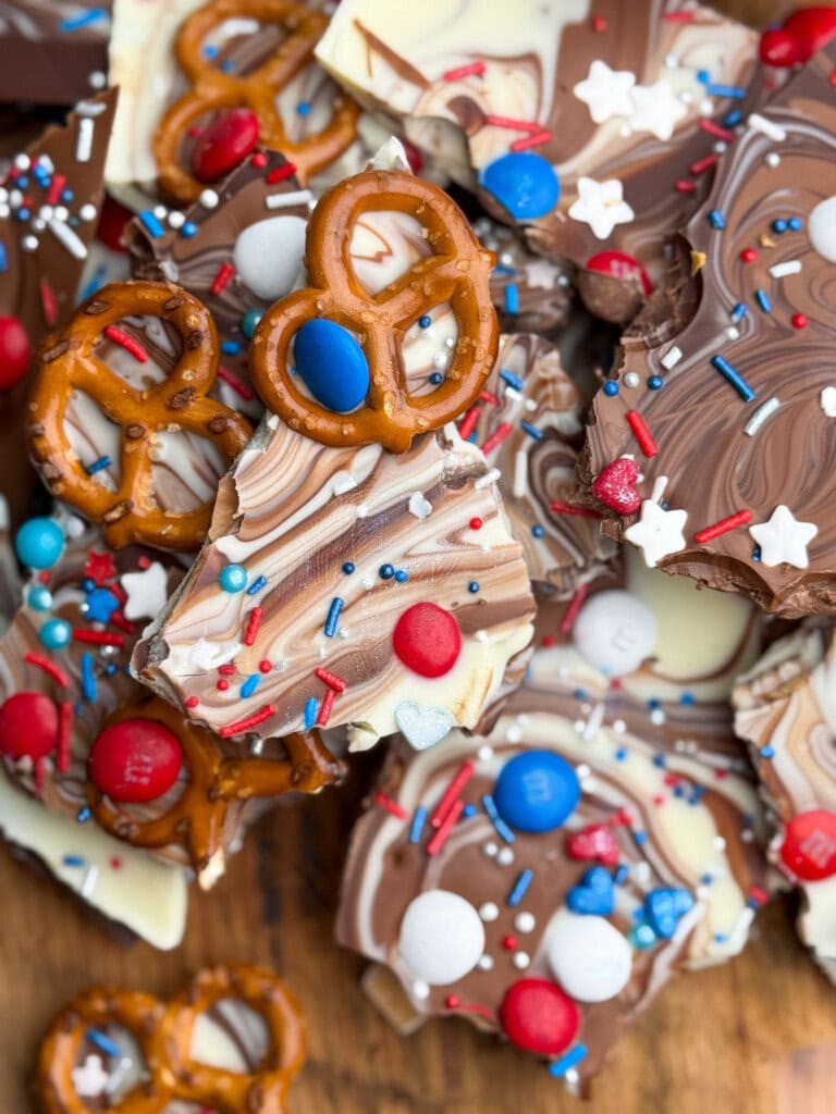 A close-up of chocolate bark pieces swirled with white and milk chocolate, topped with pretzels, red, white, and blue candies, star-shaped sprinkles arranged on a wooden surface.