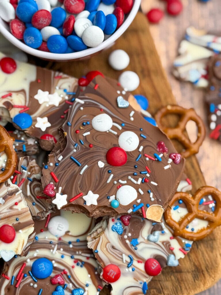 A close-up of chocolate bark swirled with milk and white chocolate, topped with red, white, and blue candies, sprinkles, and small pretzels, on a wooden board. A bowl of more red, white, and blue candies is nearby.