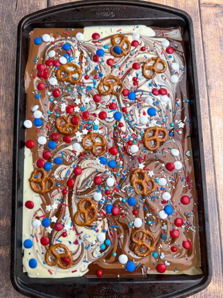 A baking sheet with swirled milk and white chocolate, topped with pretzels, red, white, and blue candy-coated chocolates, and patriotic sprinkles, on a wooden surface.