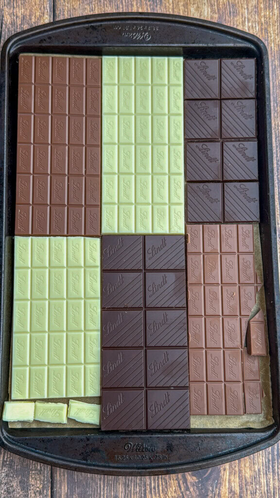 A baking tray with neatly arranged Lindt chocolate bars in three varieties: dark, milk, and white chocolate, each unwrapped and placed side by side, filling most of the tray’s surface.