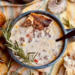 A bowl of creamy soup with ground meat, corn, and diced bell peppers, garnished with a sprig of rosemary and crusty bread. A spoon, salt and pepper shakers, and more bread are nearby on a striped cloth.