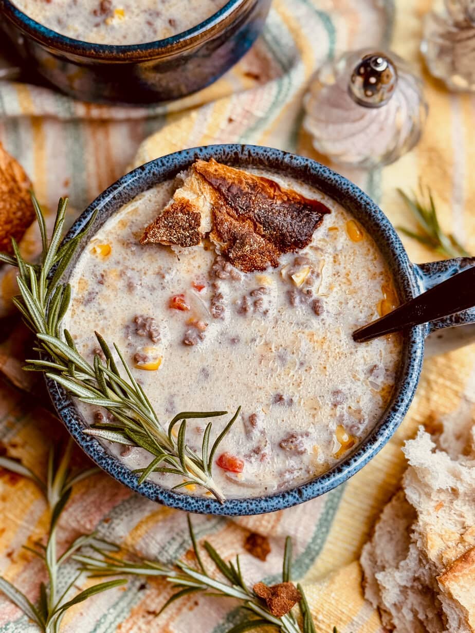 A bowl of creamy soup with ground meat, corn, and diced bell peppers, garnished with a sprig of rosemary and crusty bread. A spoon, salt and pepper shakers, and more bread are nearby on a striped cloth.